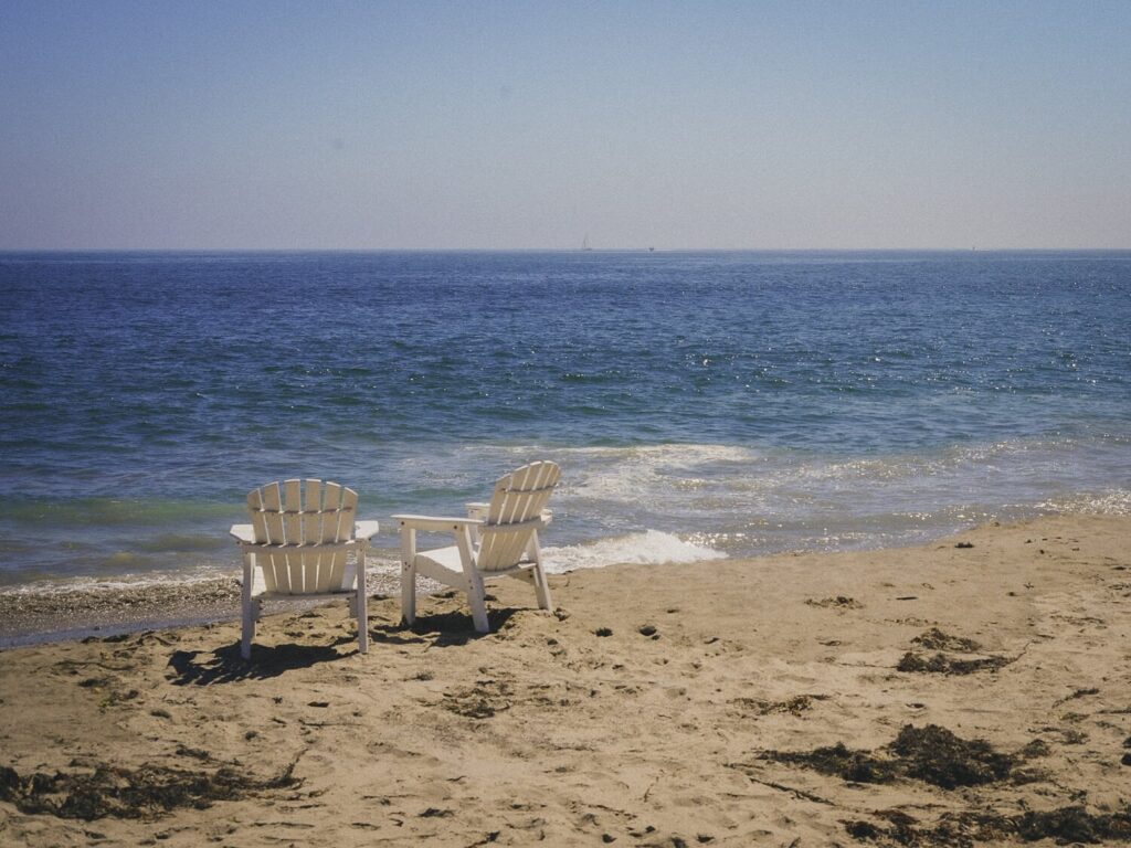 White Adirondack chairs facing the ocean on a sandy Santa Barbara beach.